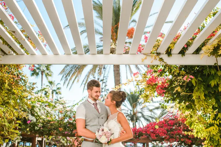 Couple standing beneath a white pergola draped in bright pink bougainvillea