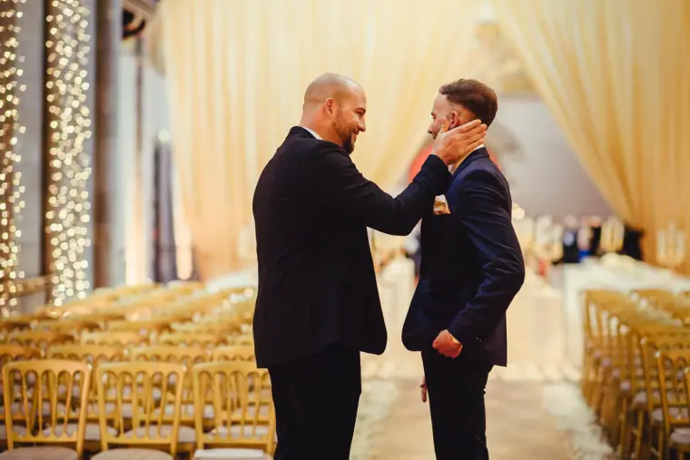 Groomsman straightening the groom's tie in the candlelit ceremony hall