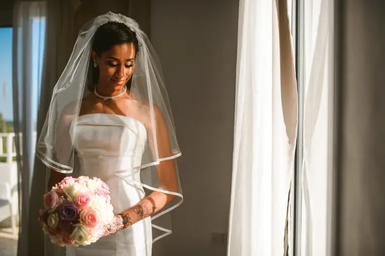 Bride holding a pastel rose bouquet beside a curtained window, veil over her shoulders