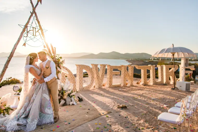 Kenton and Hassiba embracing beside giant letters and a tipi arch above the sea at Experimental Beach