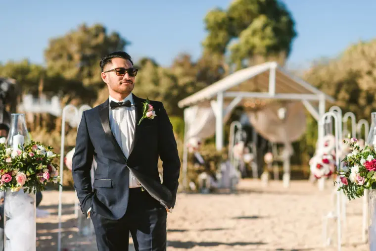 Groom in a tuxedo and sunglasses waiting by the flower-lined aisle and gazebo