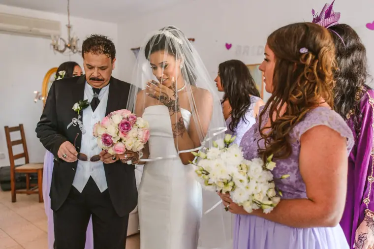 Bride in her veil wiping a tear beside her father as bridesmaids in lilac look on