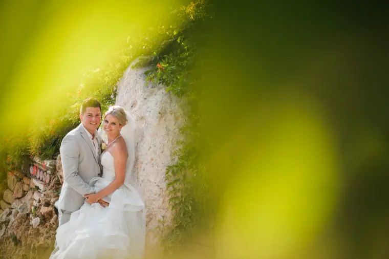 Charlotte and Nathan embracing beside a rocky wall, framed by green leaves