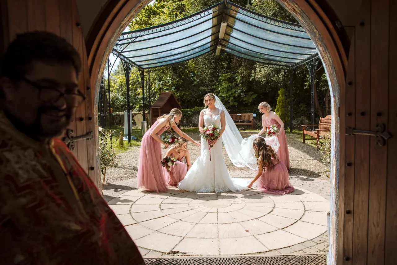 Bridesmaids in pink arranging the bride's gown in a courtyard, seen through an arched doorway