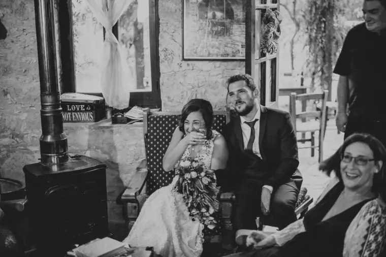 Black-and-white photo of the bride laughing beside the groom and guests by a wood stove