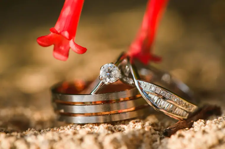 Wedding and engagement rings resting on sand beside red decorations