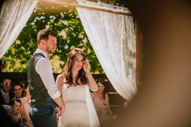 Claire wiping a tear beside Sean beneath the lace arch