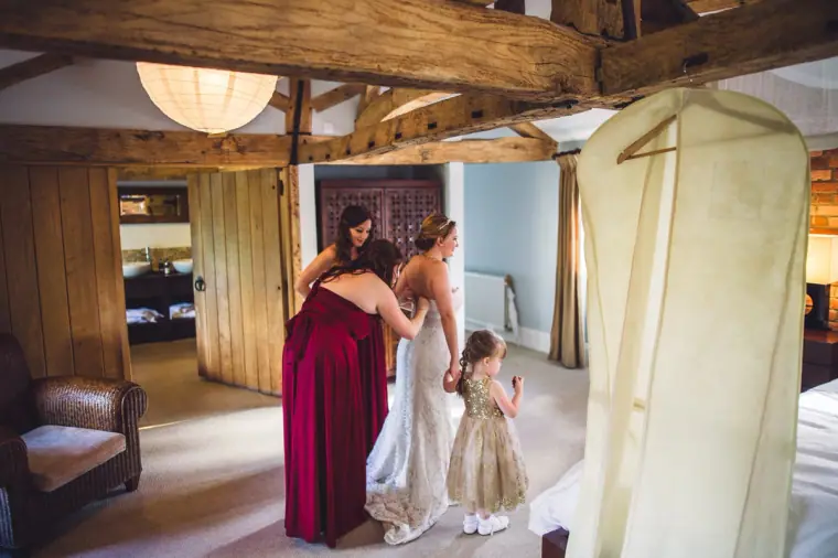 Bridesmaid in red helping Sarah into her dress as the flower girl watches