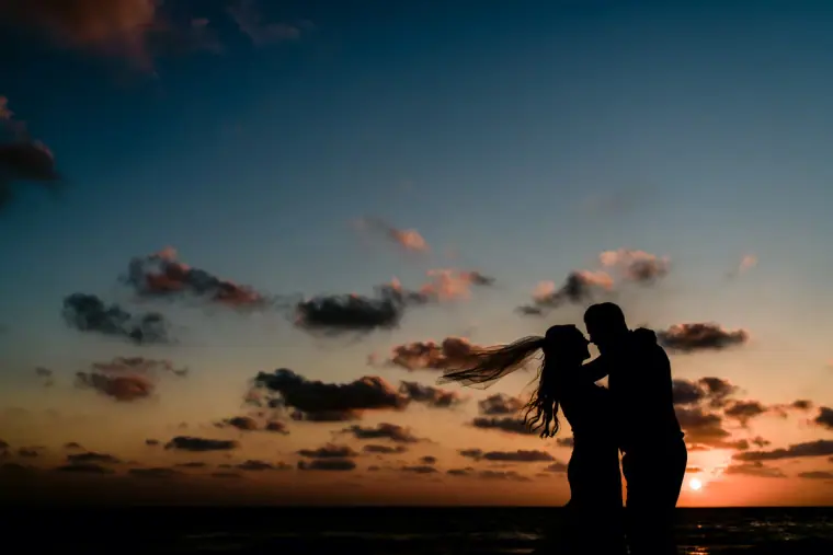 Silhouette of a couple embracing at dusk, her veil blowing above the sea