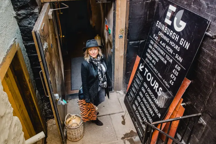 Woman in a hat smiling at the doorway of Edinburgh Gin beside a tours sign