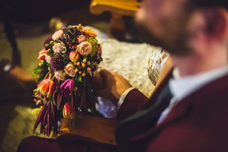 Groom in burgundy holding the bride's bouquet of roses and protea