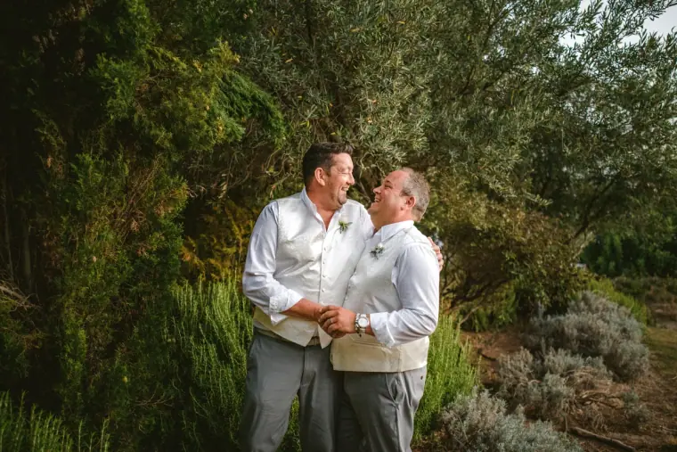Two men in white shirts and cream waistcoats laughing together among olive trees