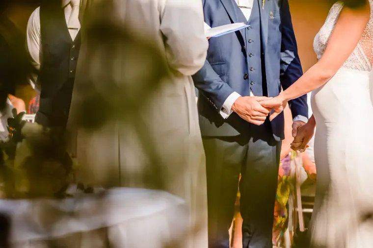 Bride and groom holding hands during the vows, glimpsed through leaves