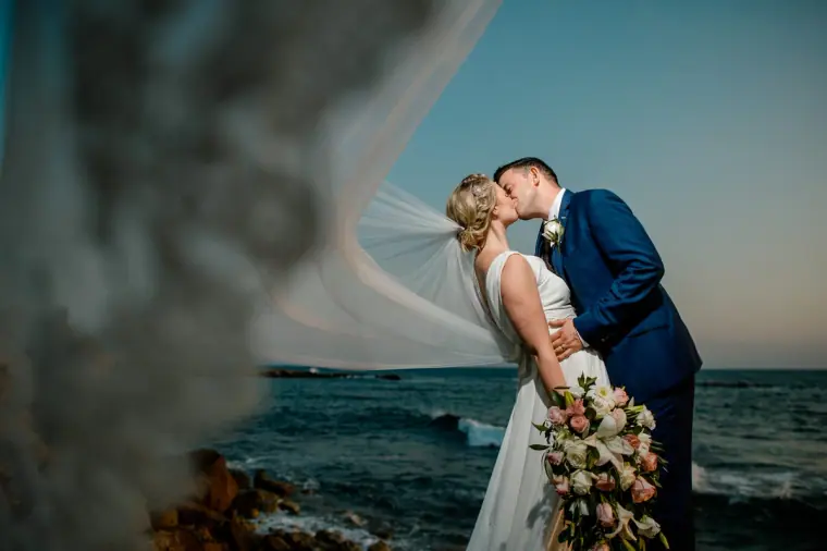 Francesca and Stephen kissing by the sea, framed by a blurred veil