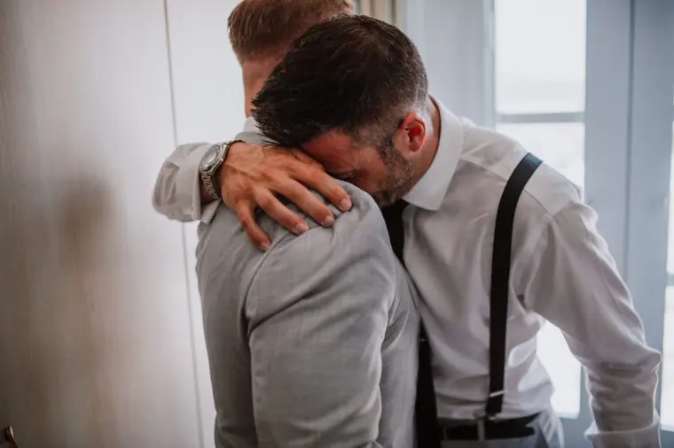 Emotional groom resting his head on a friend's shoulder in an embrace, Santorini