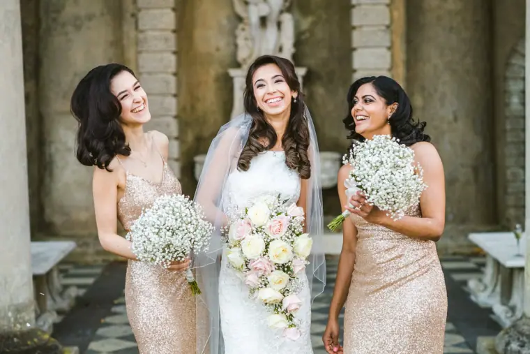Elina laughing with two bridesmaids under a stone colonnade, all holding bouquets