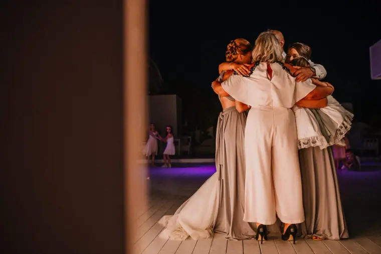 Bridesmaids and a guest in a white jumpsuit sharing a group hug, seen through a doorway