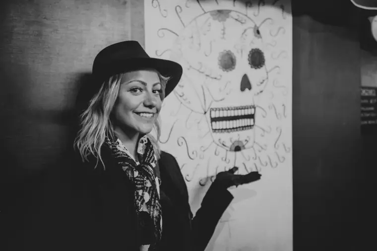 Black-and-white portrait of a smiling woman in a hat beside a sugar-skull mural