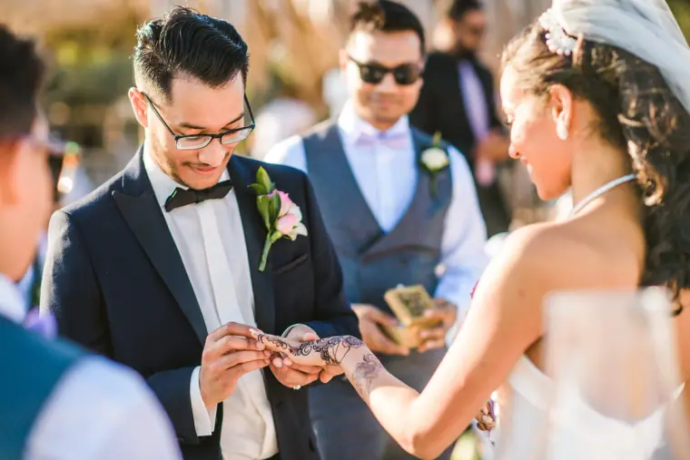 Groom slipping a ring onto the bride's hennaed hand as groomsmen look on