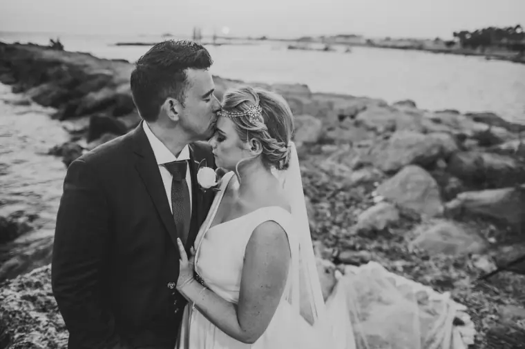 Black-and-white photo of the groom kissing the bride's forehead on the rocky shore