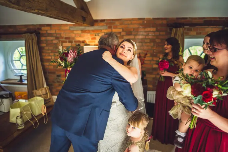 Man in a navy suit hugging the bride as bridesmaids and a toddler look on