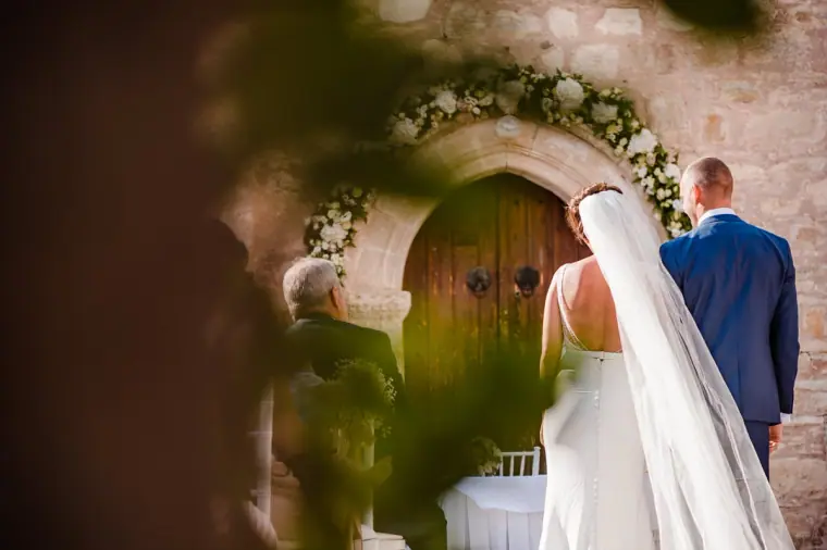 Bride and groom at the floral-framed chapel door, seen through foliage