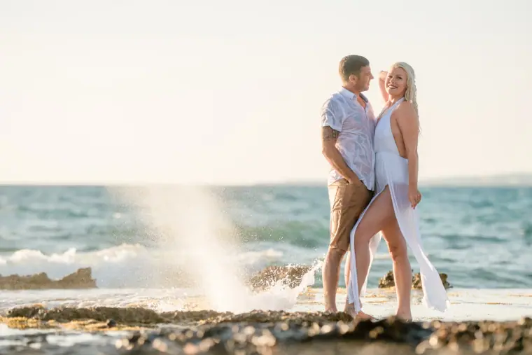 Couple standing on a rock ledge as a wave sprays up behind them