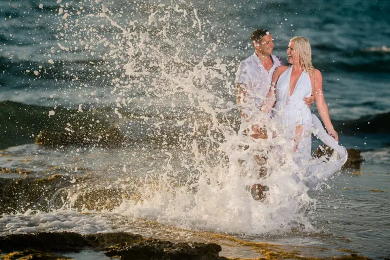 Maryellen and Ben standing on rocks as a wave crashes up in front of them, Paphos