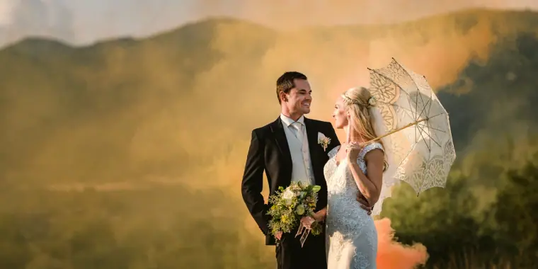 Bride with a lace parasol and groom among orange smoke above the hills