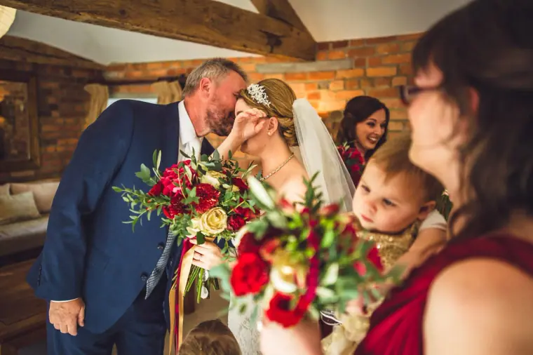 Man kissing the bride's forehead as a bridesmaid holds a red bouquet
