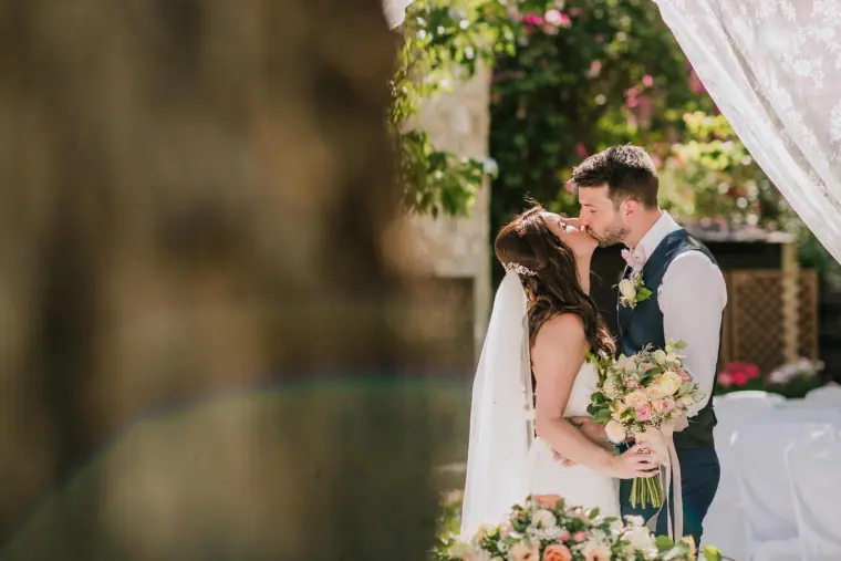 Newlyweds kissing with her bouquet in hand, glimpsed through foliage