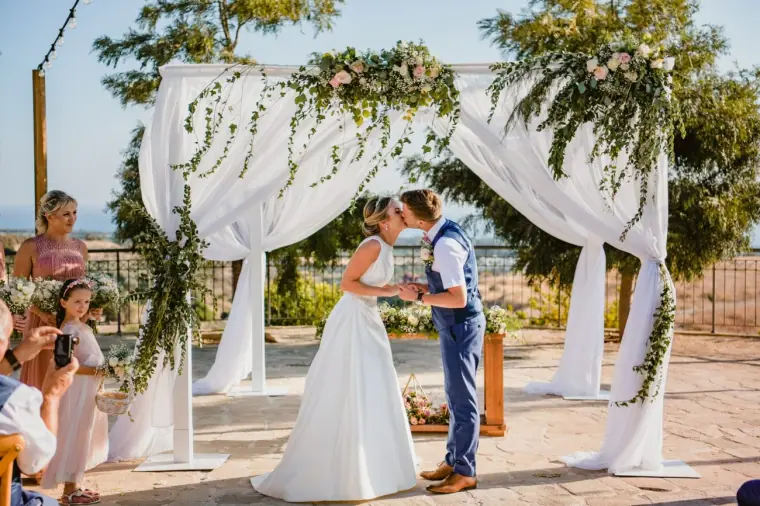 Nicola and Joel kissing beneath a draped floral arch as guests look on at Liopetro, Kouklia