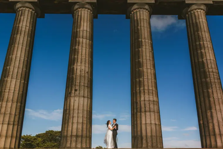 Tiny couple framed between massive stone columns under a blue sky in Scotland