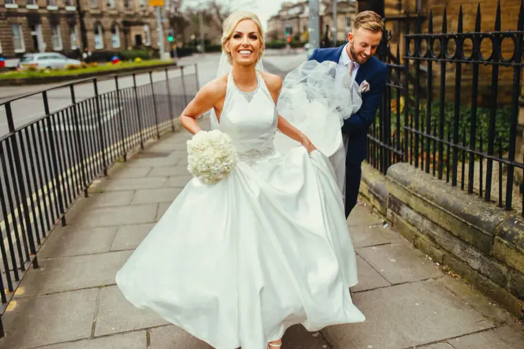 Bride laughing as she lifts her skirt on an Edinburgh pavement, groom carrying her veil