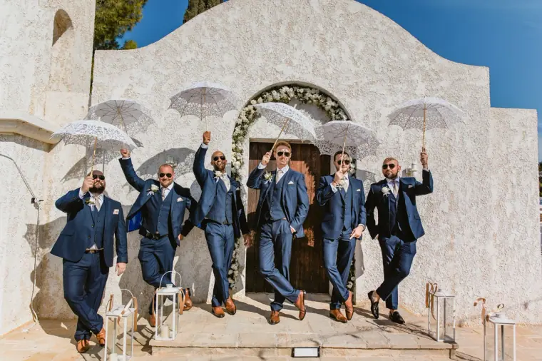 Groomsmen in navy suits leaping in front of a white chapel holding parasols