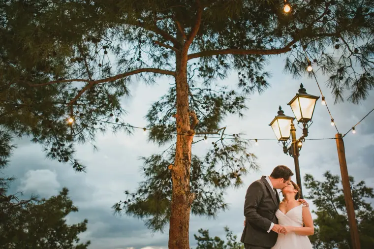 Couple embracing by a vintage lamp post under festoon lights and tall pines at dusk