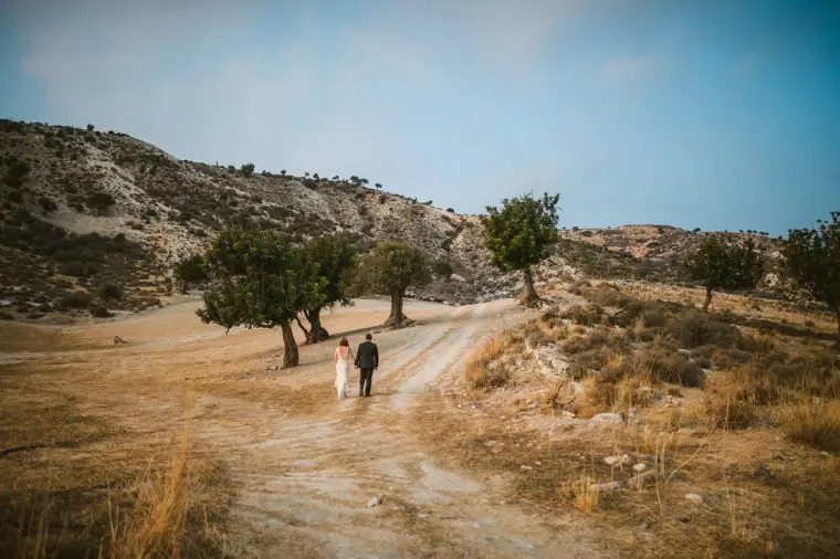 Bride and groom walking down a winding track through dry hills and carob trees