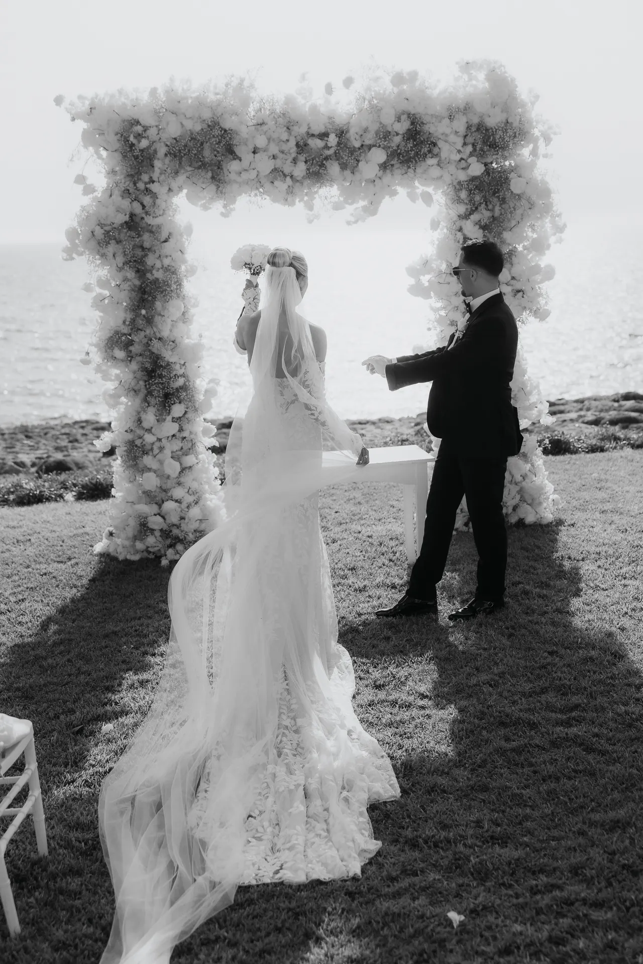 Black-and-white photo of the bride and groom facing each other beneath the floral arch