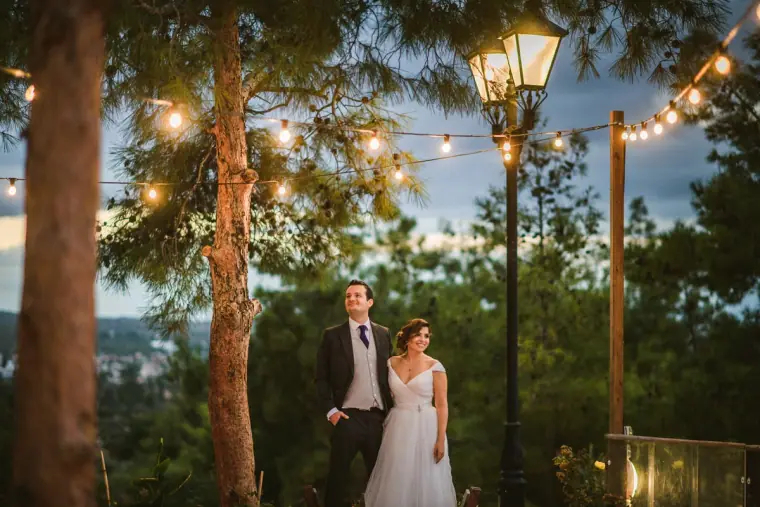 Bride and groom standing under strings of lights among tall pine trees at dusk