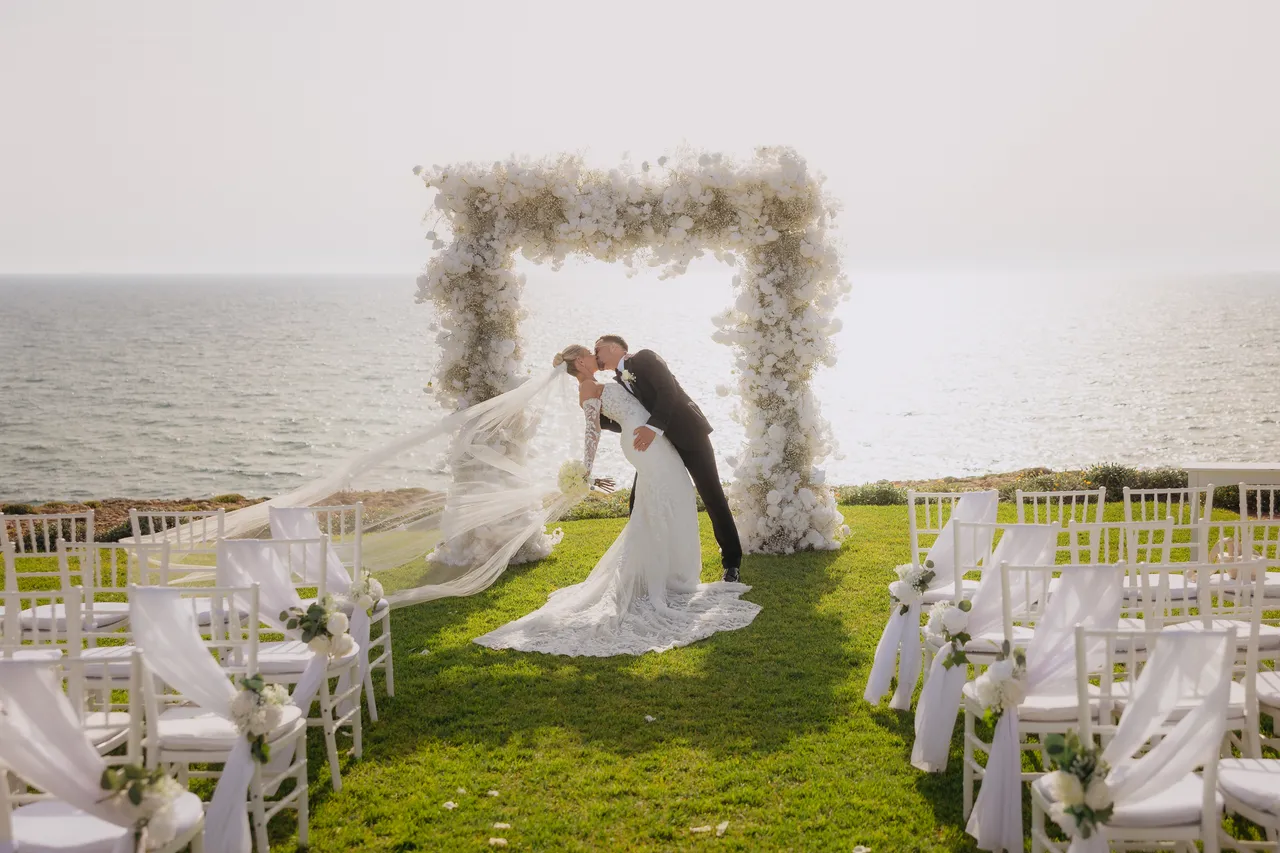 Groom dipping his bride for a kiss in front of the floral arch and the sea