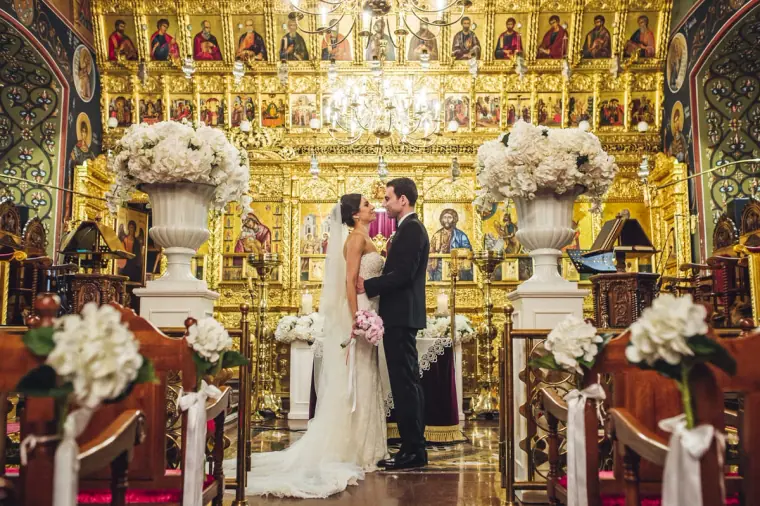 Ruby and Angelos at the ornate golden iconostasis, framed by white hydrangea arrangements