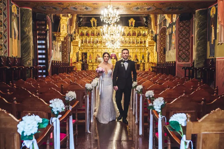 Bride and groom walking down the aisle past white floral pew ends in a gilded church