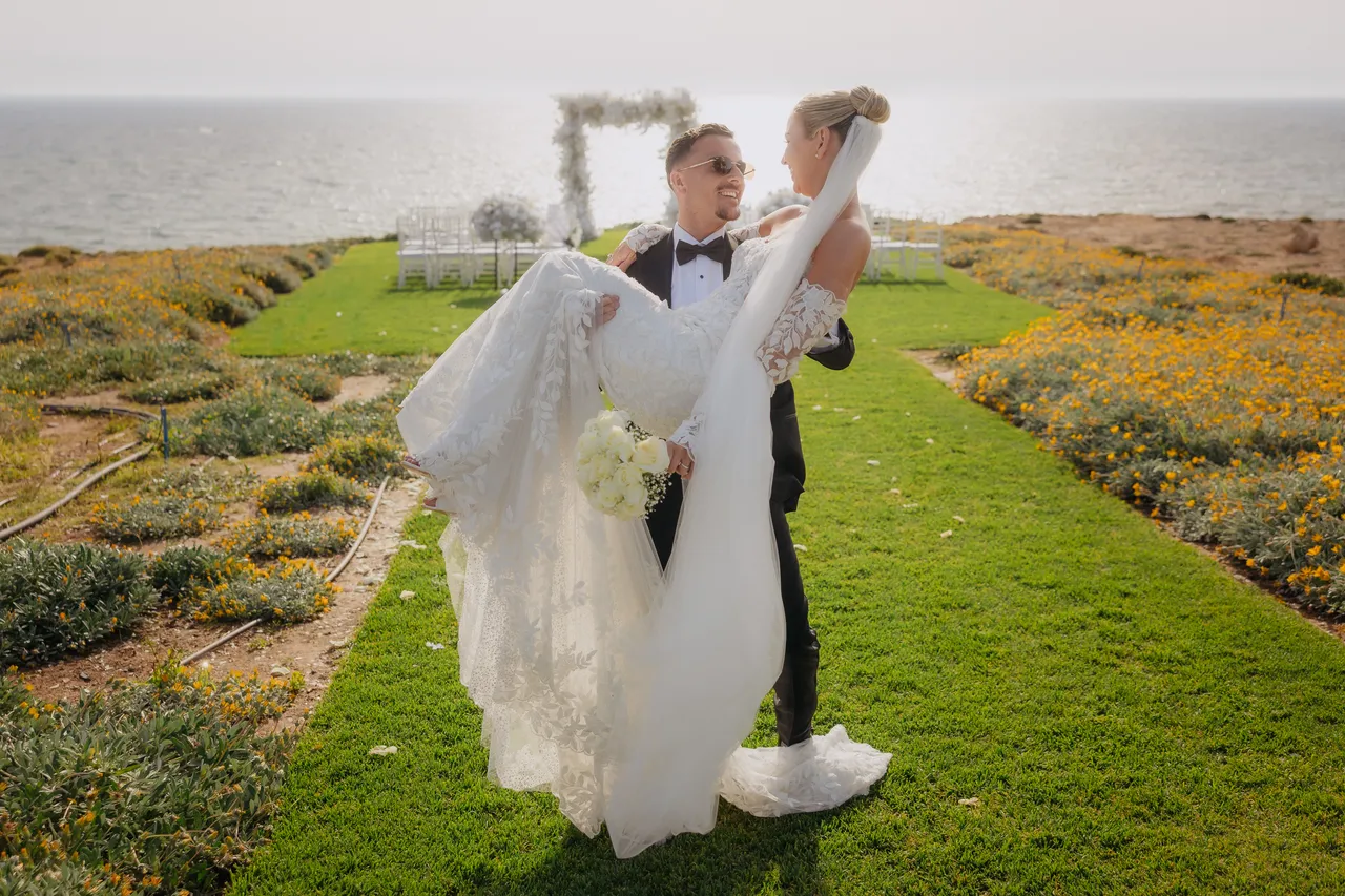 Groom carrying his bride along a grassy path with the arch and sea behind