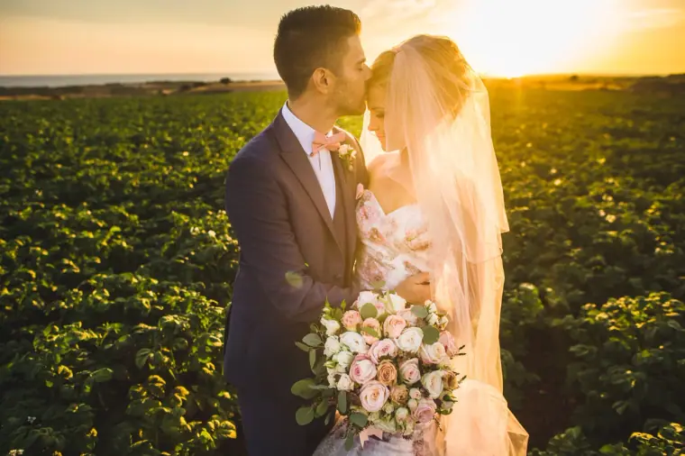 Groom kissing the bride's forehead in a field at sunset, bouquet in hand