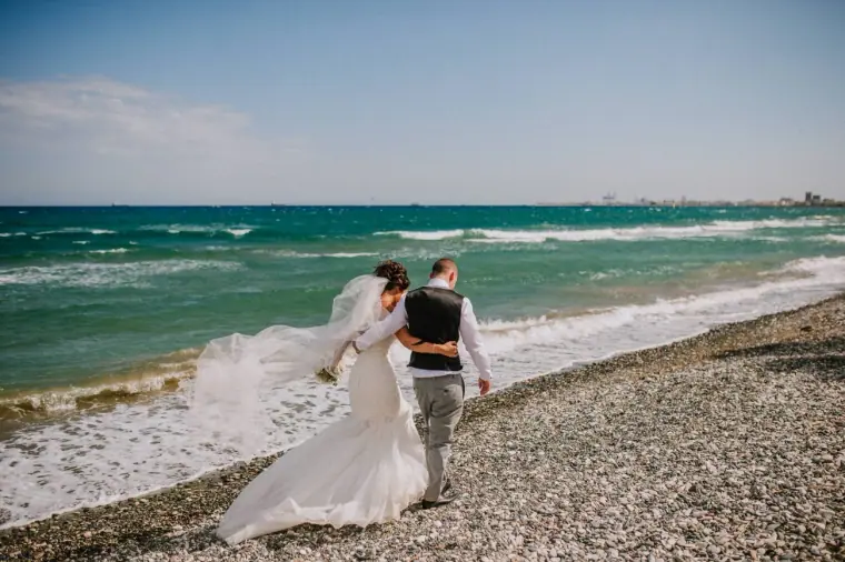 Louise and Mike walking along the shingle shore, her veil blowing, at Galu Seaside, Larnaca