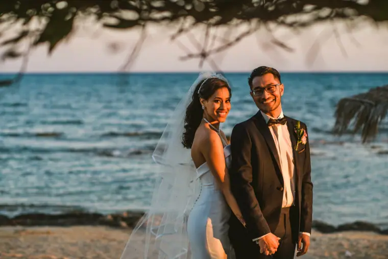 Raya and Adhib standing side by side smiling on the shoreline at golden hour