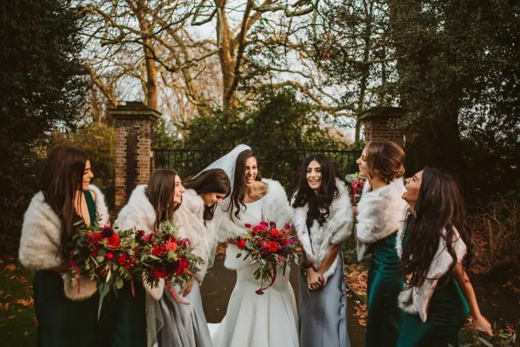 Bride laughing with bridesmaids in white faux-fur wraps holding red bouquets
