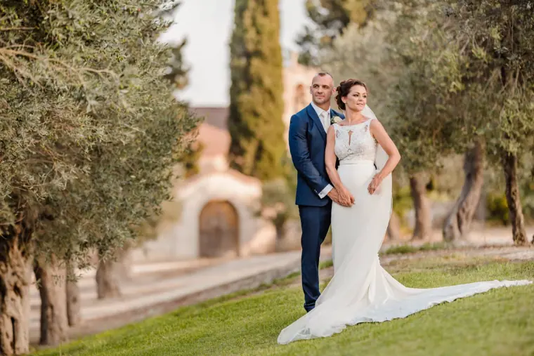 Bride in a fitted gown with a long train beside the groom among olive trees
