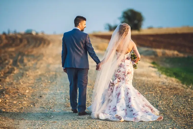 Bride and groom walking hand in hand down a dirt track, her floral train trailing