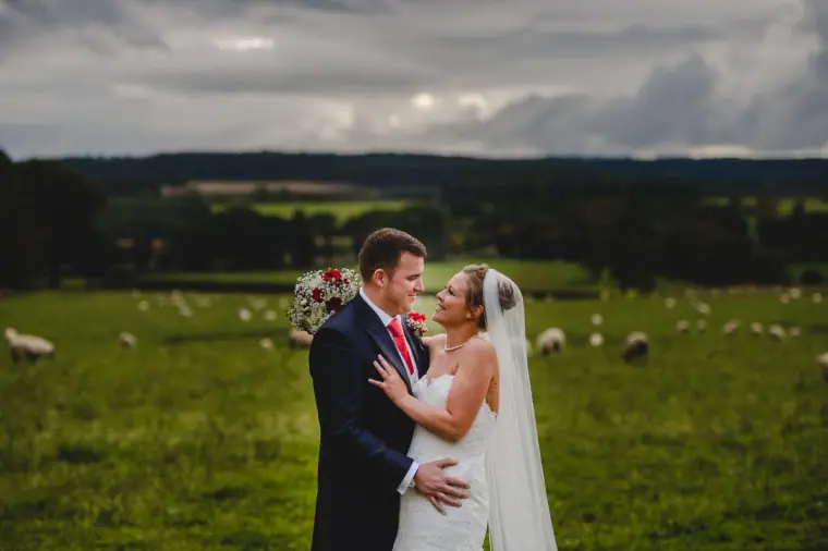 Sonny and Jo embracing in a field of grazing sheep with Northumberland hills beyond